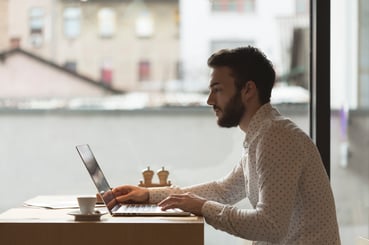 Jeune homme barbu concentré travaillant sur un ordinateur portable prêt d'une baie vitrée lumineuse, tasse de café posée sur la table, illustrant le travail en freelance