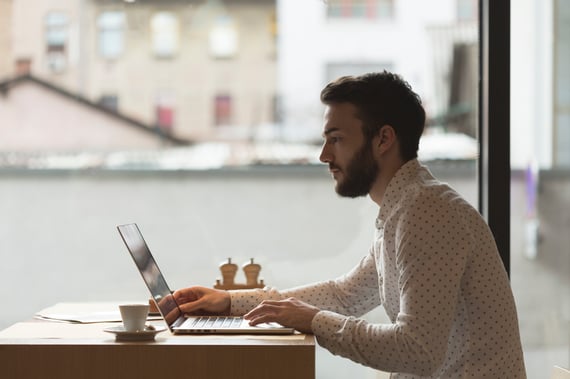 Jeune homme barbu concentré travaillant sur un ordinateur portable prêt d'une baie vitrée lumineuse, tasse de café posée sur la table, illustrant le travail en freelance
