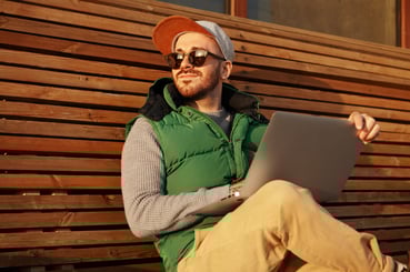 Homme barbu assis sur un banc en bois, portant une casquette, des lunettes de soleil, un pull gris et une doudoune verte, travaillant sur un ordinateur portable tout en profitant du soleil.