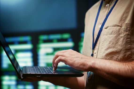 Technicien en informatique utilisant un ordinateur portable devant un écran affichant des données système ou réseau, illustrant un environnement de supervision ou d'administration système.