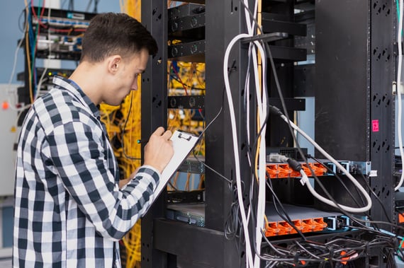 Technicien réseau inspectant une baie de serveurs et prenant des notes sur un clipboard, entouré de câbles et d’équipements informatiques.