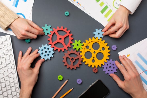 Plusieurs personnes assemblent des engrenages colorés symbolisant le travail d’équipe, la collaboration et l’optimisation des processus, avec des graphiques et un clavier visibles sur la table.